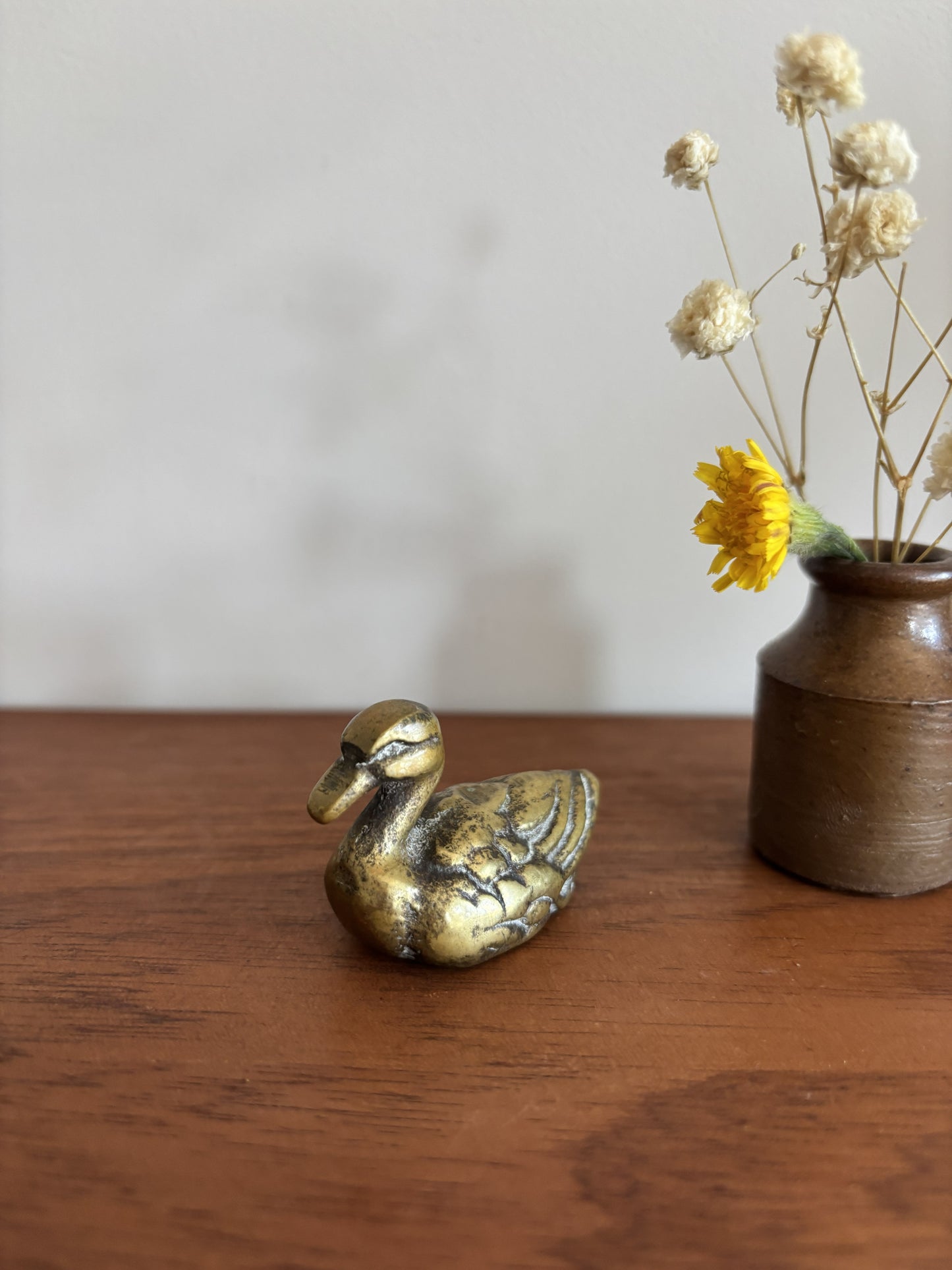 A small brass duck figurine next to a rustic brown ceramic vase with dried white flowers and a yellow accent flower, placed on a wooden table against a plain light gray wall.