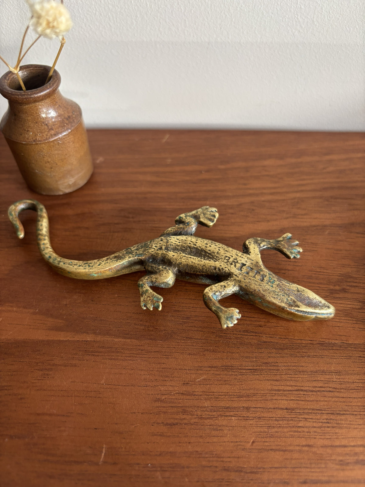 A small brass lizard figurine resting on a wooden tabletop, with a small ceramic vase containing dried stems in the background.