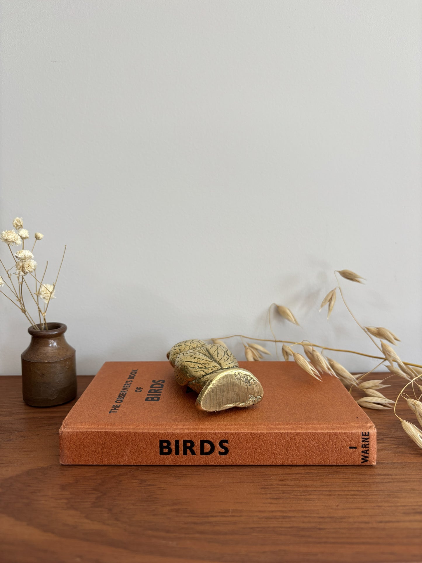 The base of a brass owl figurine on a brown book titled 'BIRDS' on a wooden table, styled with a small vase of dried flowers and a decorative branch.
