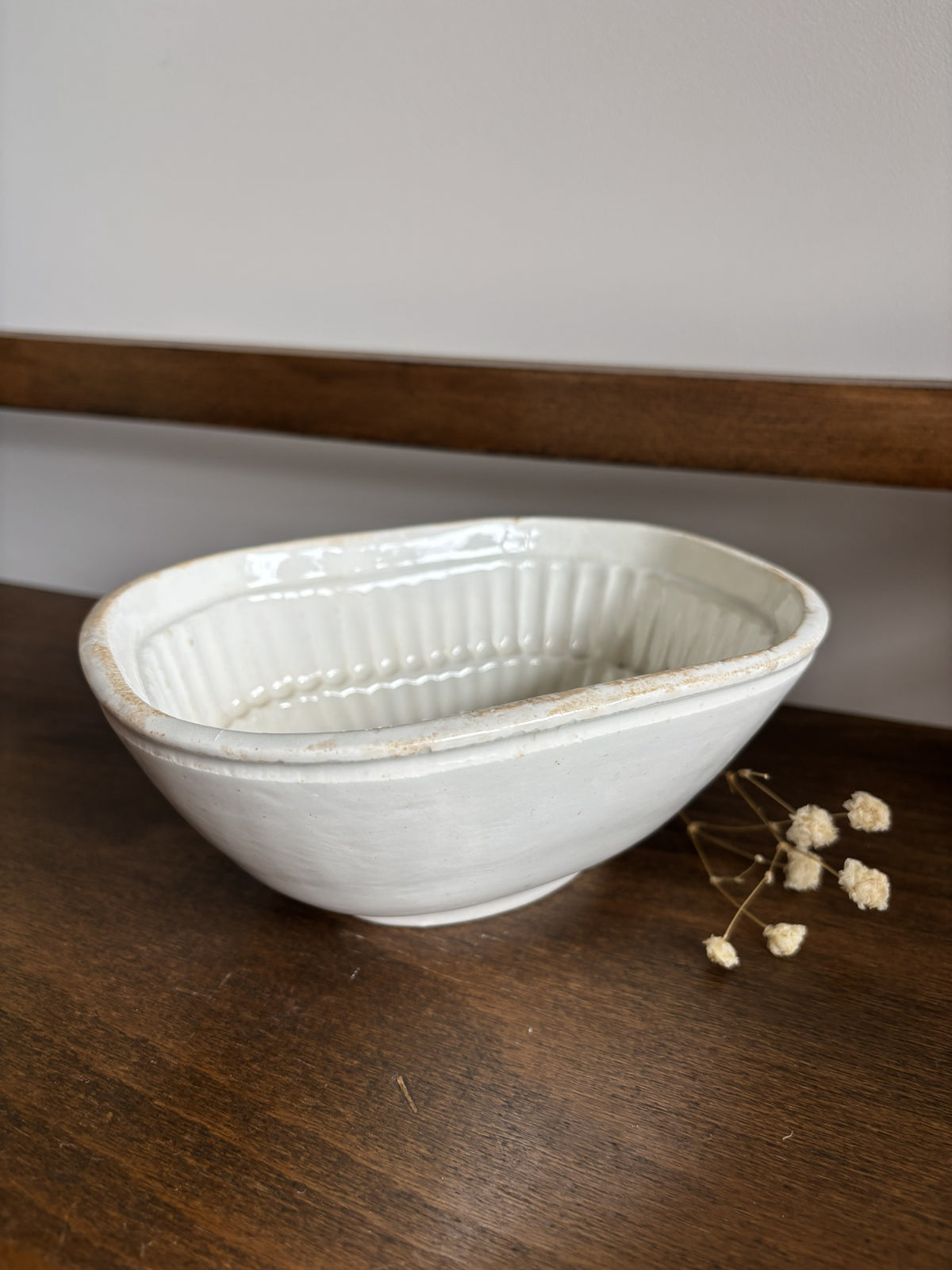A white ceramic jelly mould with a ribbed interior, resting on a dark wooden surface next to small dried flowers.