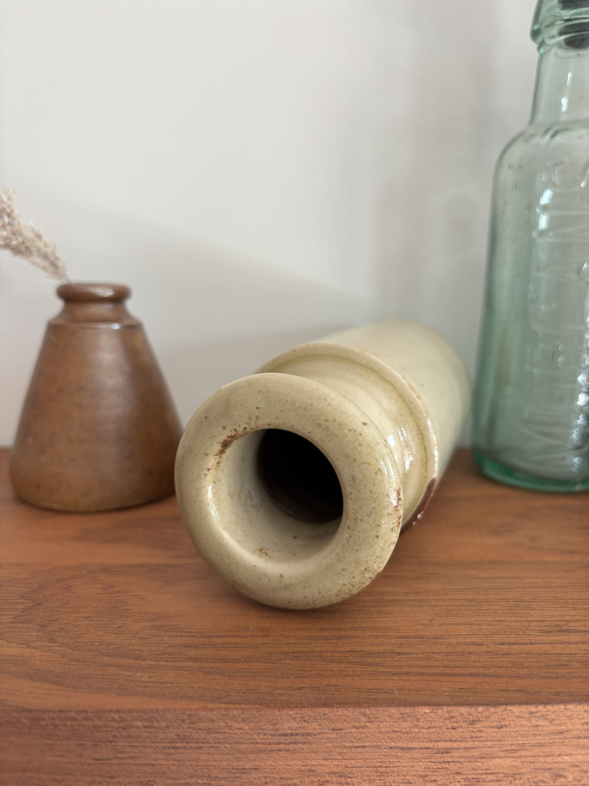 A close up of the top of a vintage cream stoneware ink bottle displayed on a wooden shelf against a plain light gray wall styled with a vase with dried grass and a tall translucent green glass bottle
