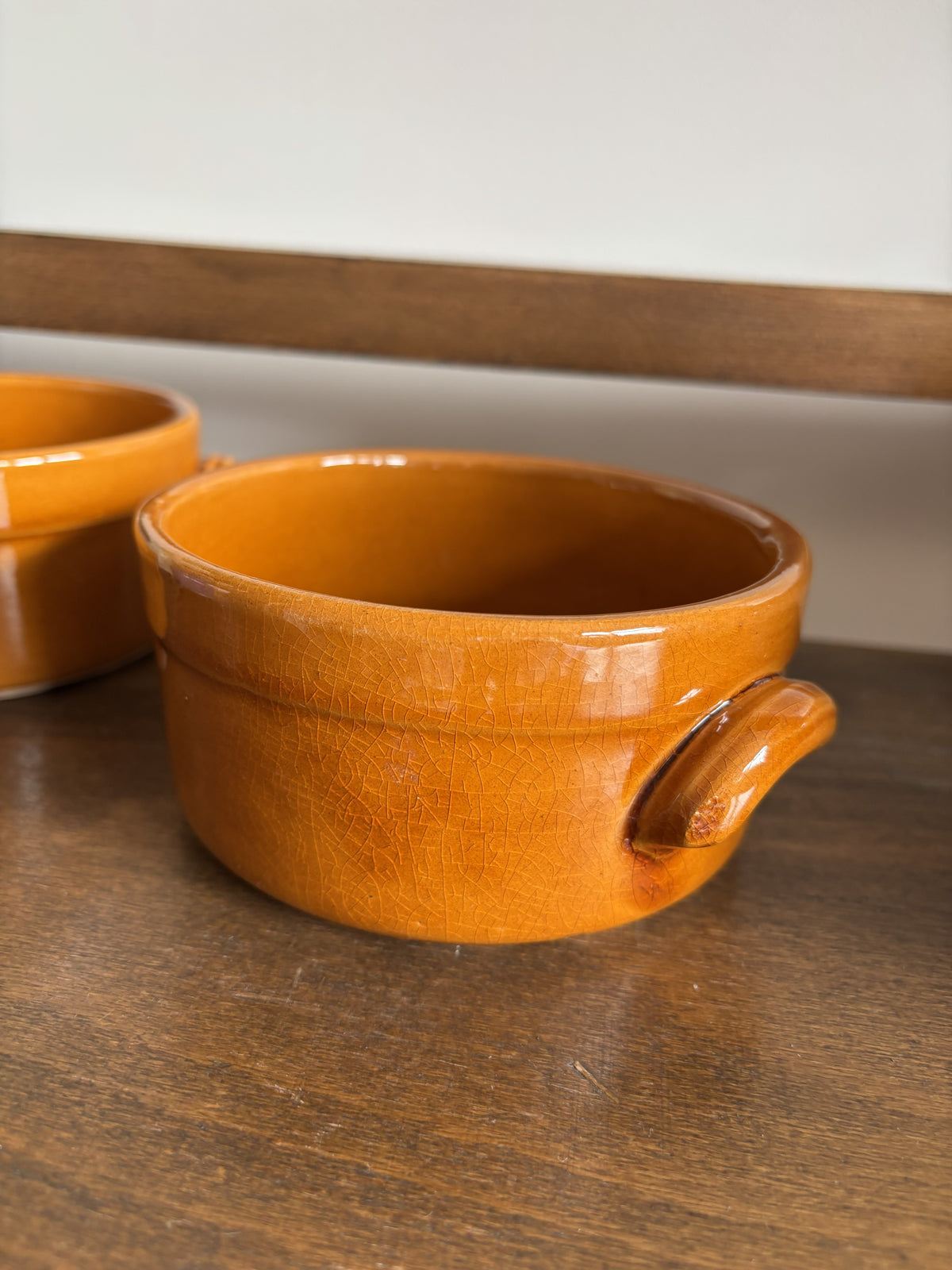A small orange glazed ceramic bowl with a side handle, shown on a dark wooden surface with another similar bowl partially visible in the background.