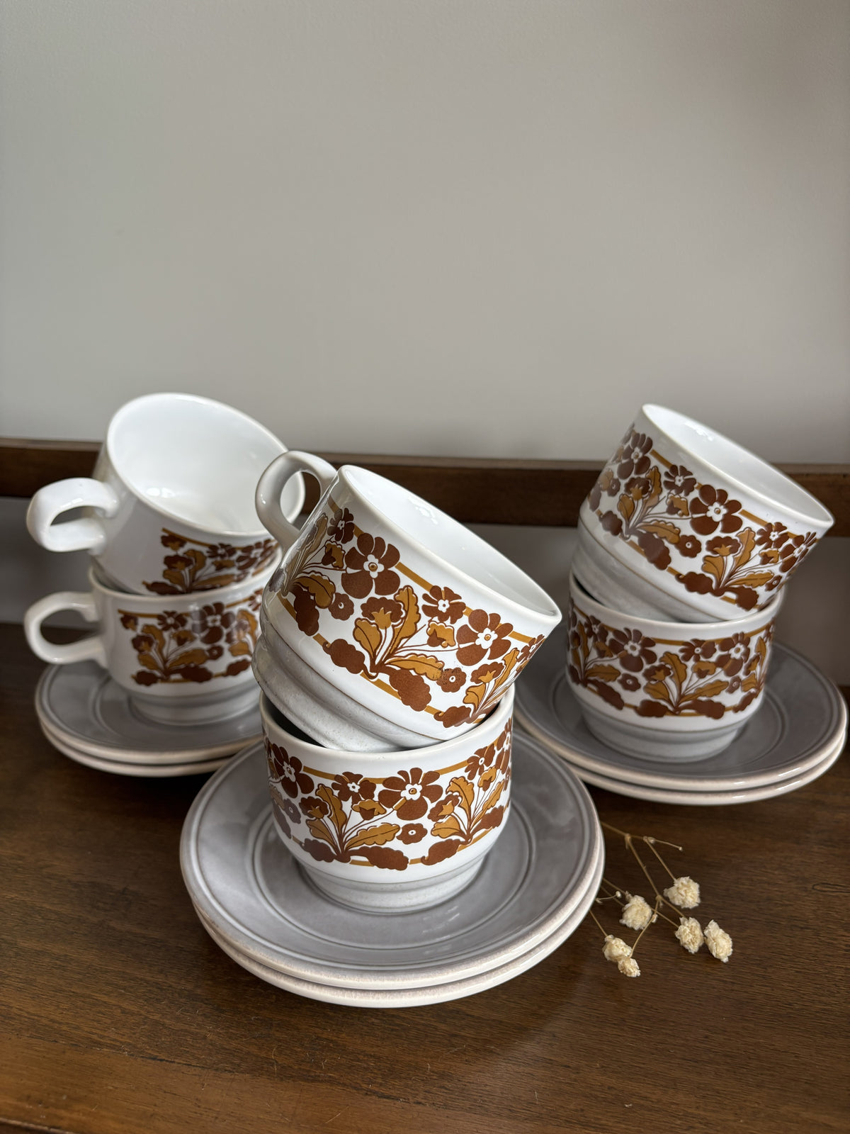 A set of vintage white ceramic teacups with saucers, decorated with a brown botanical pattern, arranged on a wooden table with a neutral background.