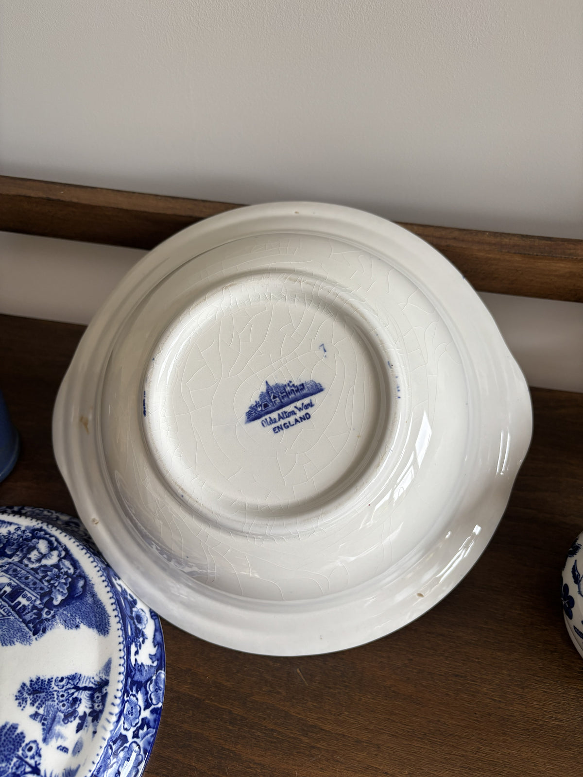 An upside-down tureen dish with a blue printed crest and text in the center, resting on a dark wooden shelf next to other blue-and-white patterned dishes. Makers mark can be seen in the centre.