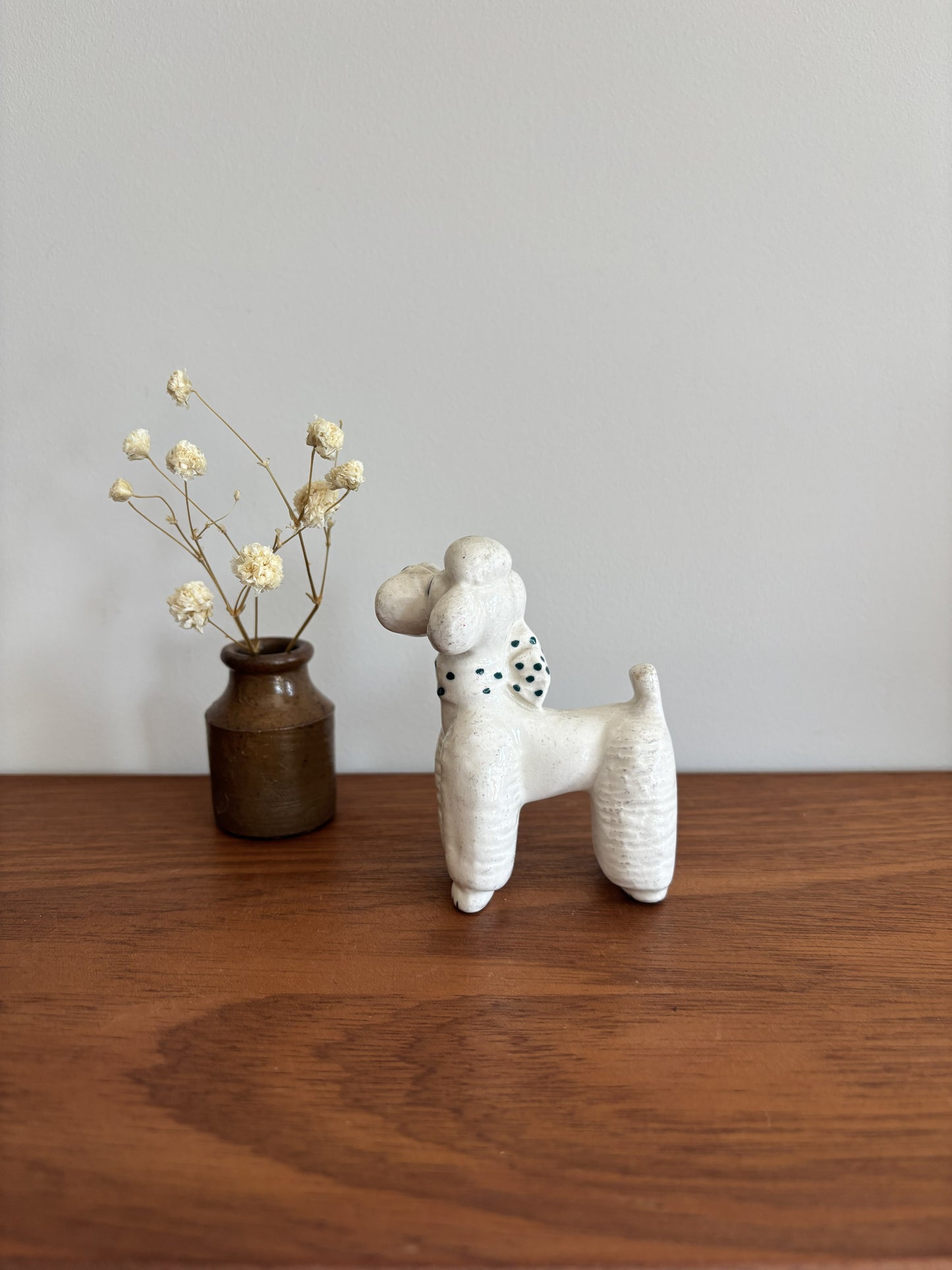 A white textured ceramic poodle figurine with green spots on its neck stands on a wooden table next to a small brown vase with dried cream flowers against a plain light gray wall.