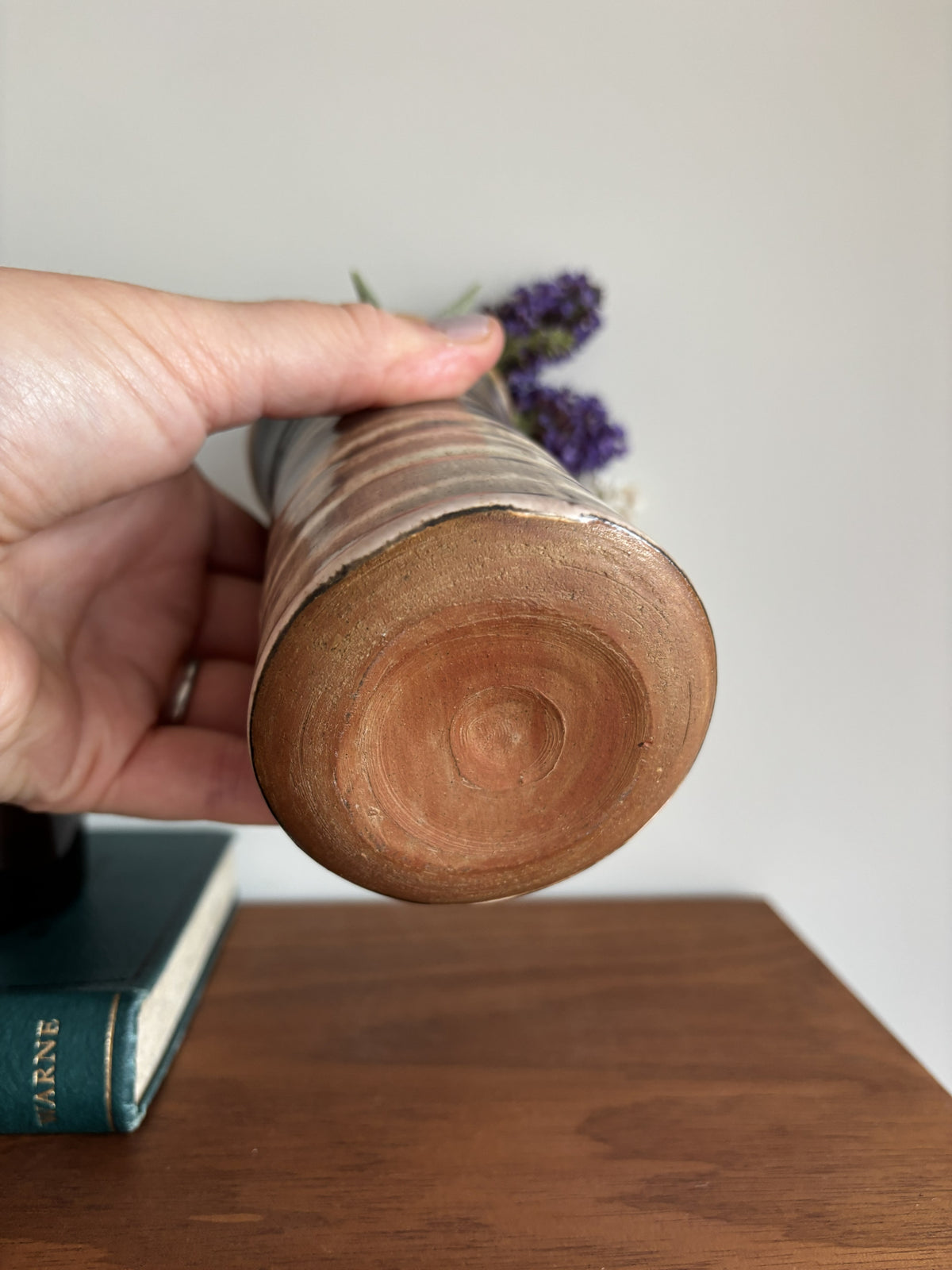 A hand holds a handmade ceramic vase with a rustic wood-grain base and a narrow metallic silver neck, containing dried purple flowers, set on a wooden table.