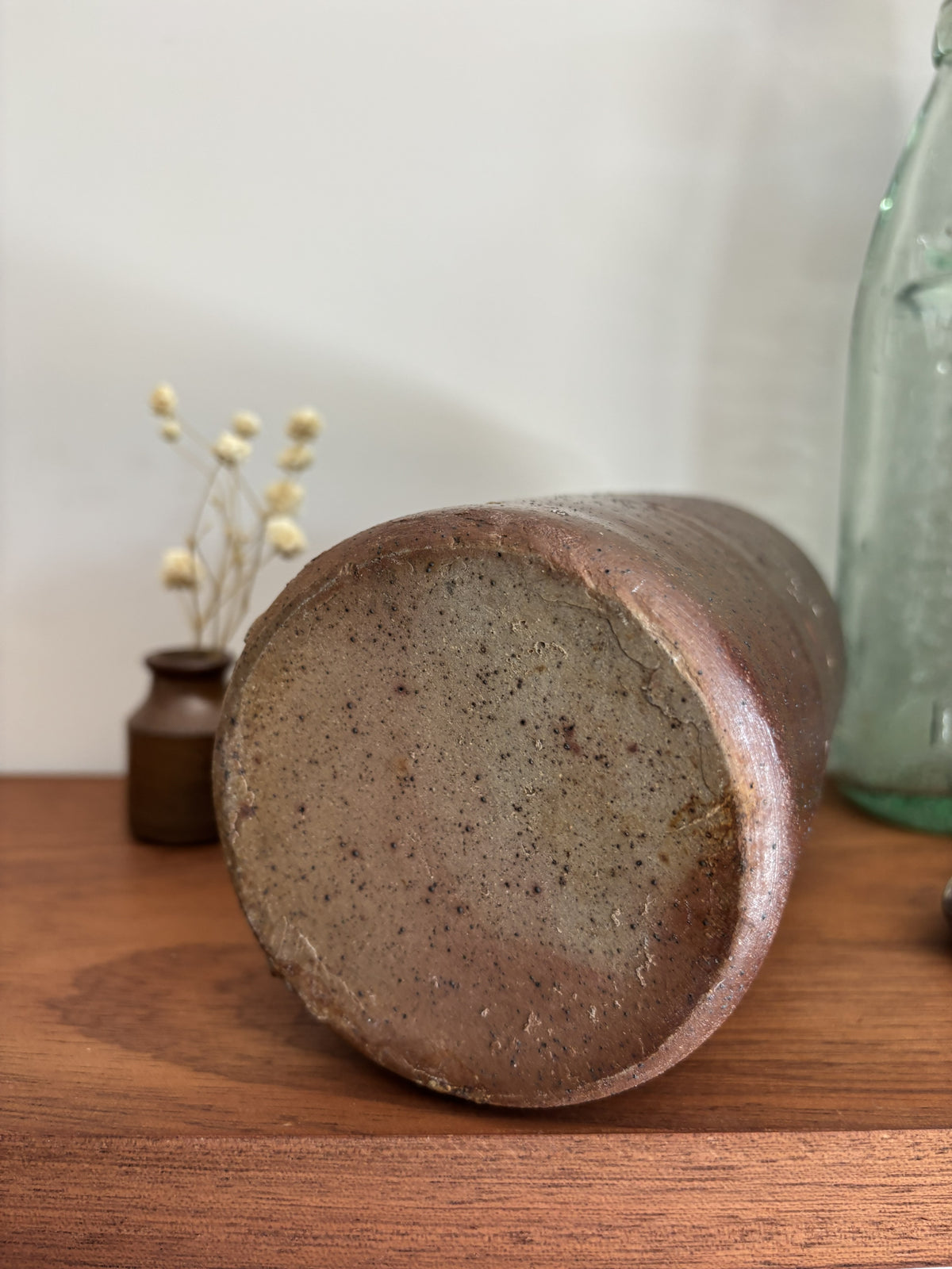 The base of a vintage brown stoneware bottle with a textured, speckled glaze stands on a wooden table against a plain light gray wall. To its left is a small vase with dried white flowers, and to its right is a vintage embossed light green glass bottle with a silver decorative object in front.