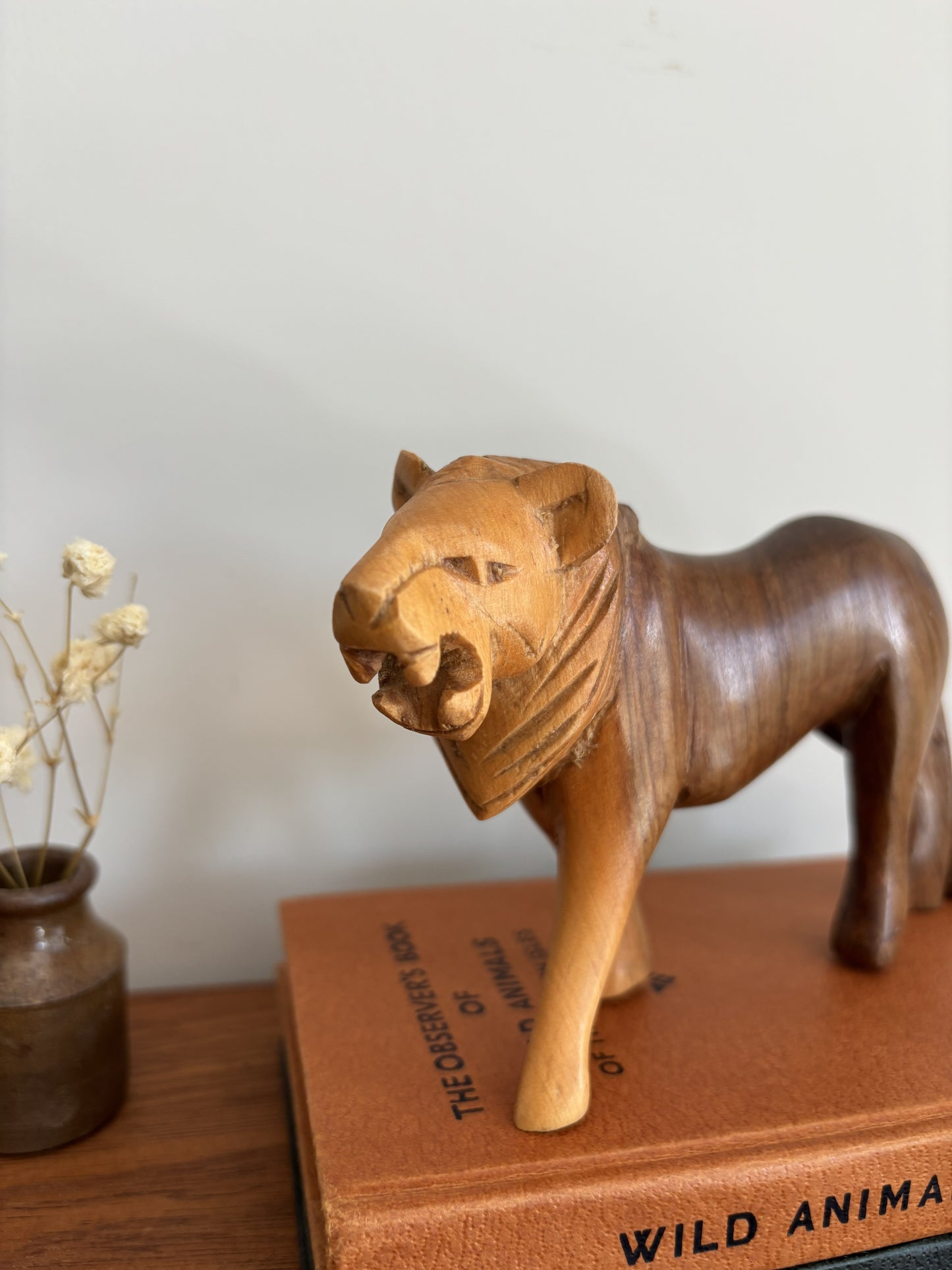A hand-carved wooden lion figurine with a natural light-to-dark brown gradient, displayed on top of a vintage brown book next to a small vase of dried flowers.