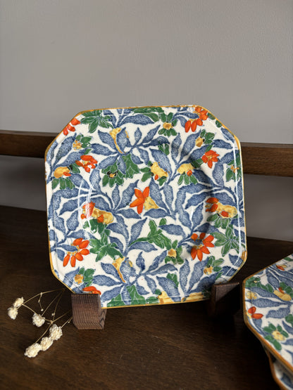 A square ceramic plate with rounded corners, featuring a floral pattern in blue, green, orange, and yellow on a white background, displayed on a wooden stand.