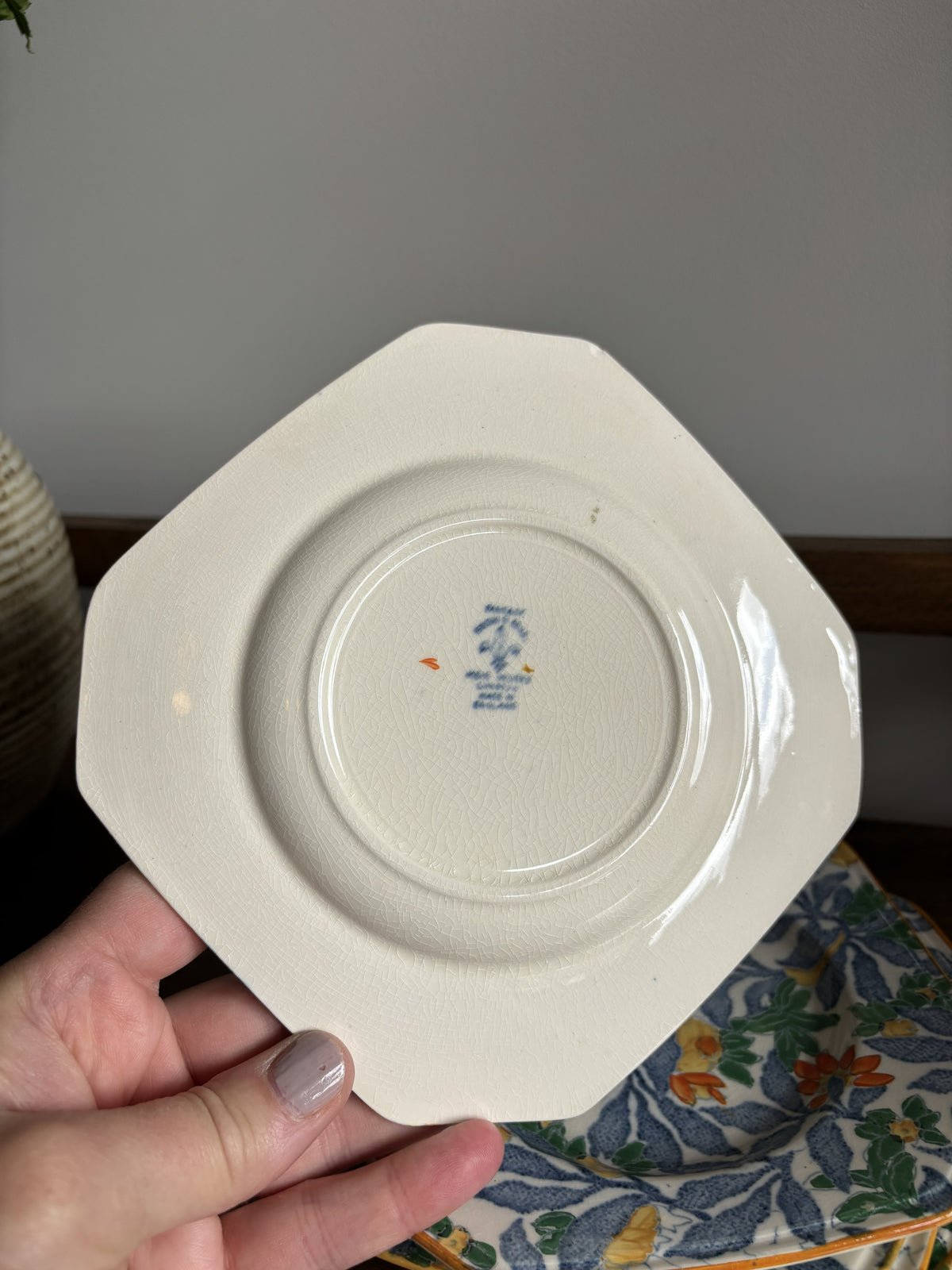 A hand holding an octagonal off-white ceramic plate or saucer, showing the bottom side with a small blue and orange printed mark in the center. The background features a gray wall, a wooden shelf, and a floral-patterned textile.