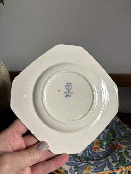 A hand holding an octagonal off-white ceramic plate or saucer, showing the bottom side with a small blue and orange printed mark in the center. The background features a gray wall, a wooden shelf, and a floral-patterned textile.
