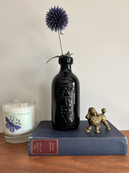 A glossy black Batey & Co glass bottle vase with embossed lettering, holding a single purple-blue globe flower, styled on a blue book next to a brass poodle figurine and a candle on a wooden surface.
