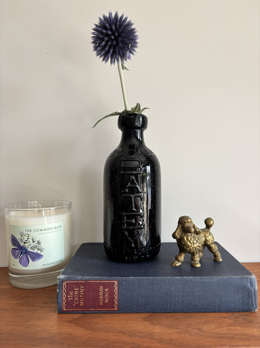 A glossy black Batey & Co glass bottle vase with embossed lettering, holding a single purple-blue globe flower, styled on a blue book next to a brass poodle figurine and a candle on a wooden surface.