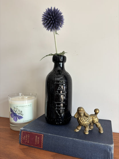 A glossy black Batey & Co glass bottle vase with embossed lettering, holding a single purple-blue globe flower, styled on a blue book next to a brass poodle figurine and a candle on a wooden surface.
