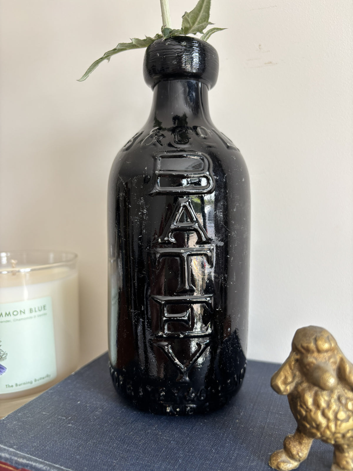 A glossy black Batey & Co glass bottle vase with embossed lettering, holding a single purple-blue globe flower, styled on a blue book next to a brass poodle figurine and a candle on a wooden surface.