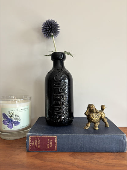 A glossy black Batey & Co glass bottle vase with embossed lettering, holding a single purple-blue globe flower, styled on a blue book next to a brass poodle figurine and a candle on a wooden surface.