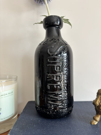 A glossy black Batey & Co glass bottle vase with embossed lettering, holding a single purple-blue globe flower, styled on a blue book next to a brass poodle figurine and a candle on a wooden surface.