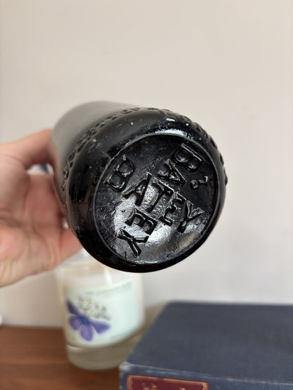 A glossy black Batey & Co glass bottle vase with embossed lettering, holding a single purple-blue globe flower, styled on a blue book next to a brass poodle figurine and a candle on a wooden surface.