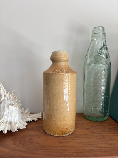 A vintage stoneware bottle with a warm beige glaze displayed on a wooden surface next to a vintage green glass bottle and a white seashell.