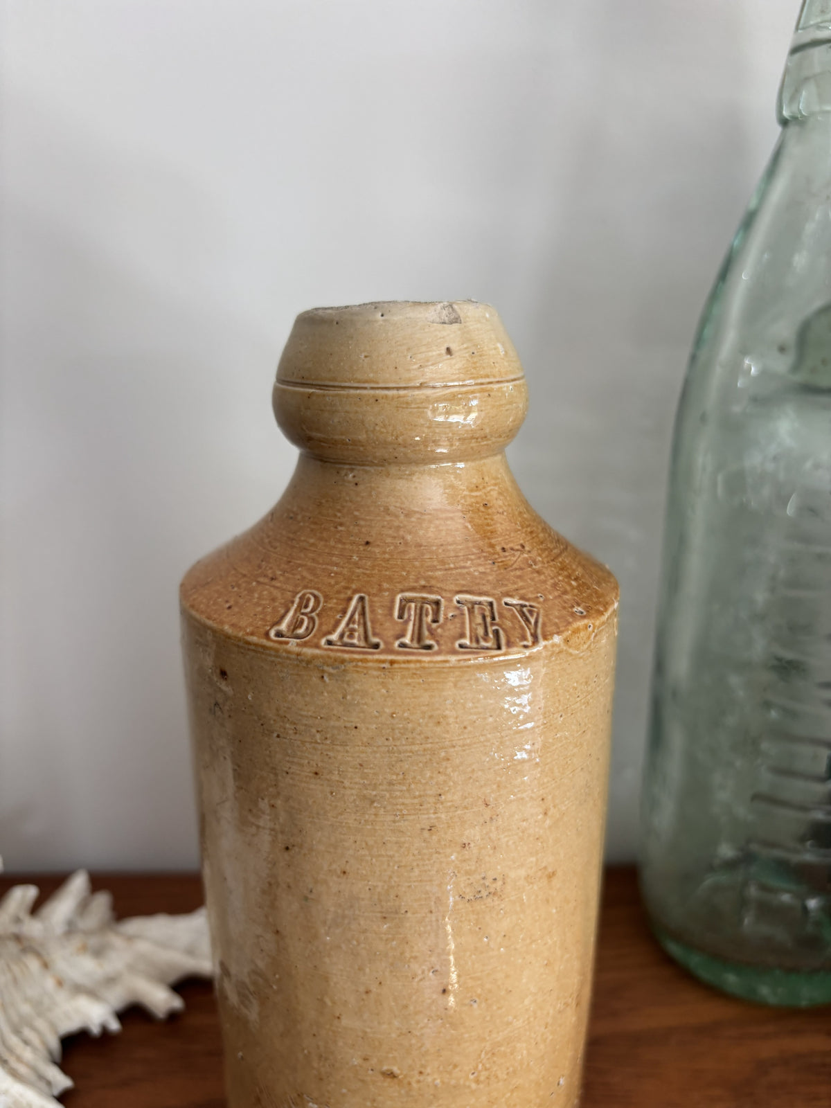 A vintage stoneware bottle with a warm beige glaze displayed on a wooden surface next to a vintage green glass bottle and a white seashell.  The word Batey is embossed on the neck. 