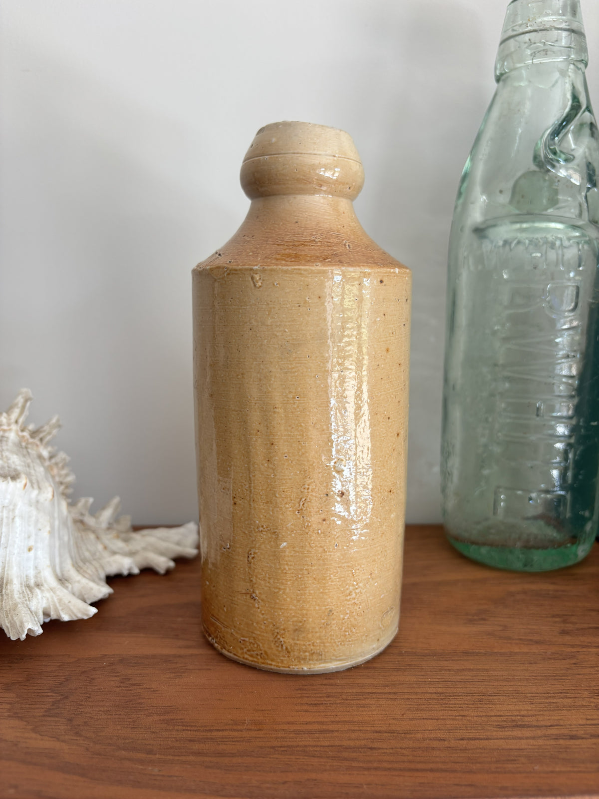 A vintage stoneware bottle with a warm beige glaze displayed on a wooden surface next to a vintage green glass bottle and a white seashell.