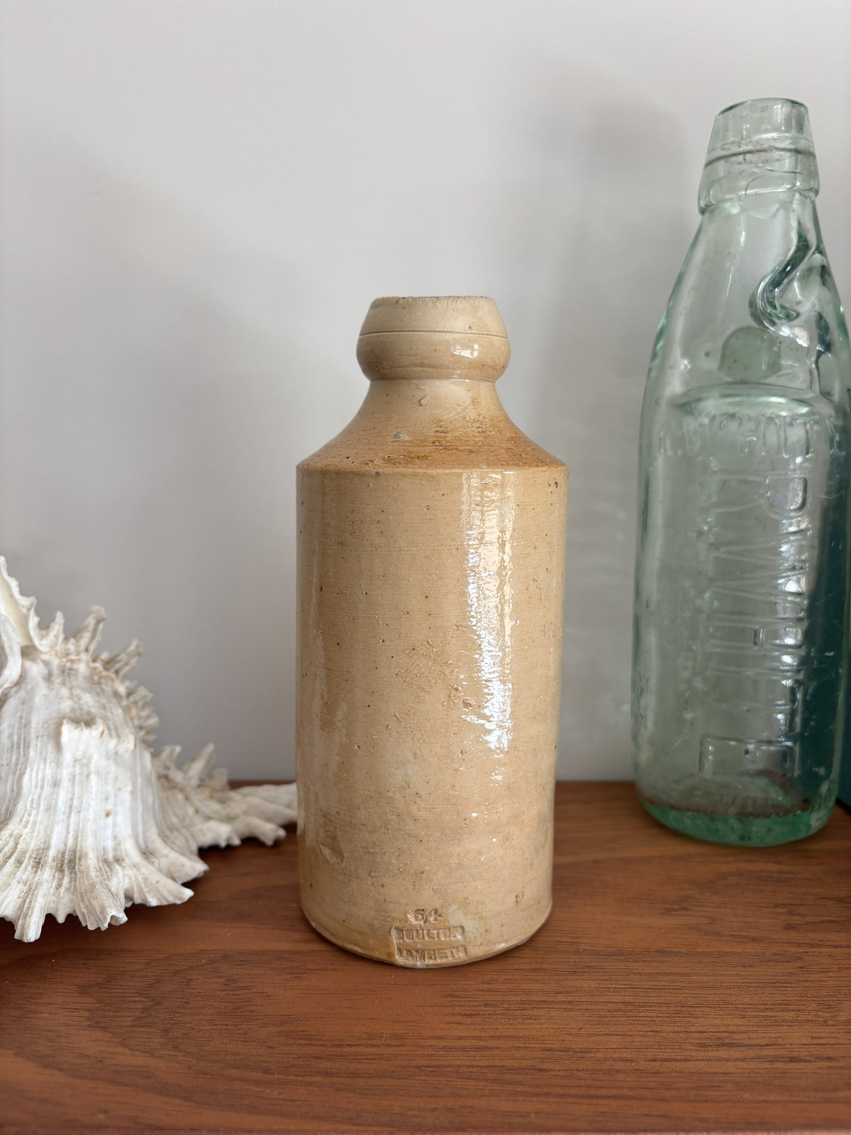 A vintage stoneware bottle with a warm beige glaze displayed on a wooden surface next to a vintage green glass bottle and a white seashell.