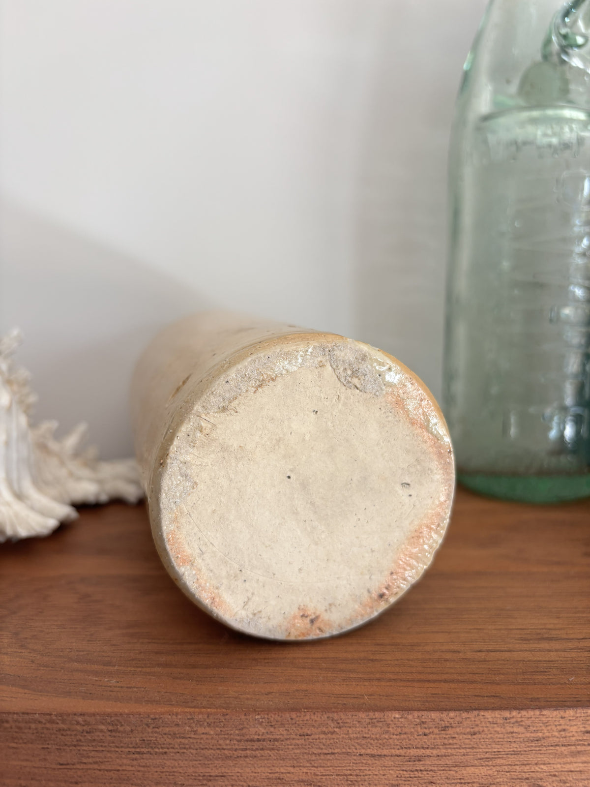 The base of a vintage stoneware bottle with a warm beige glaze displayed on a wooden surface next to a vintage green glass bottle and a white seashell.