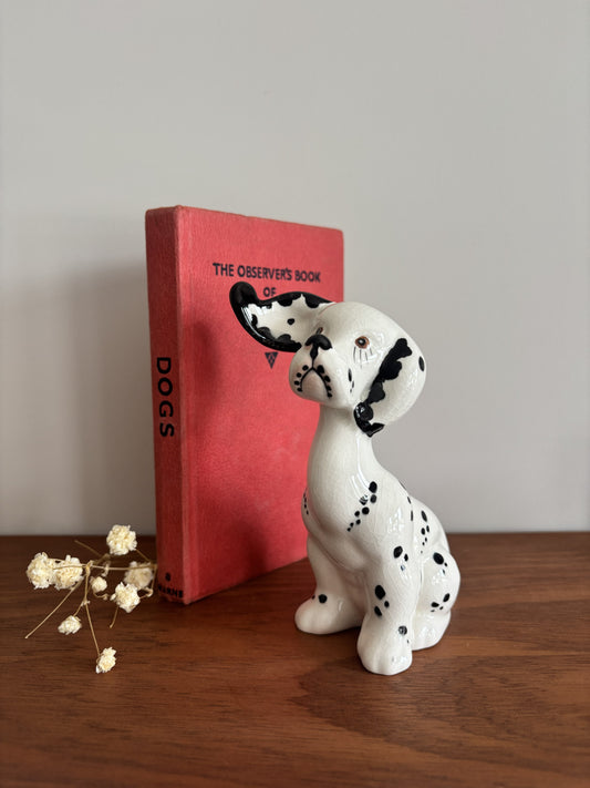 A white ceramic Dalmatian dog figurine with black spots sits on a wooden table next to a red book titled 'The Observer's Book of Dogs' and small dried white flowers against a plain light gray wall.