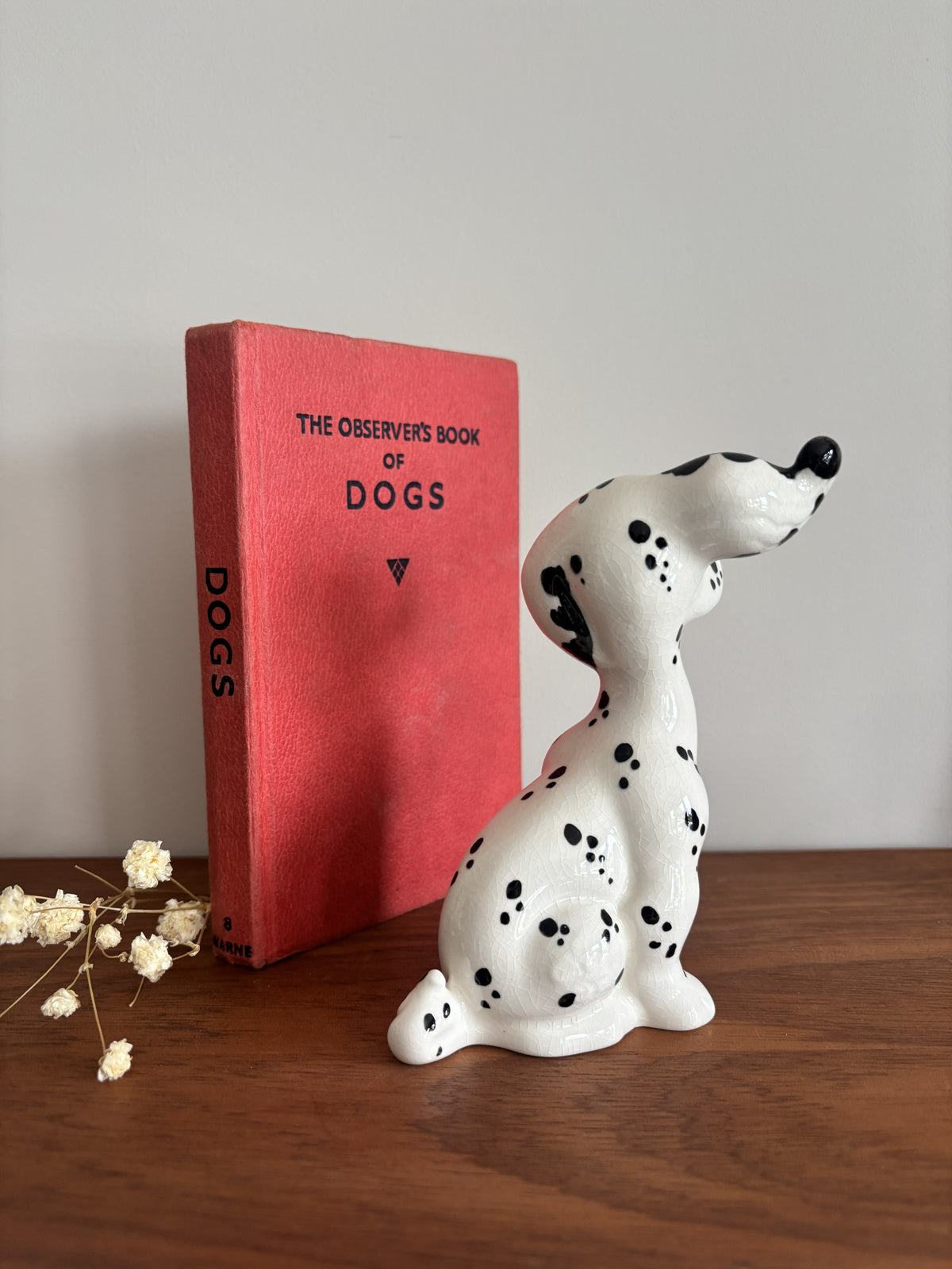 A white ceramic figurine of a Dalmatian dog with black spots sits on a wooden table next to a red hardcover book titled 'The Observer's Book of Dogs' against a plain light gray wall.