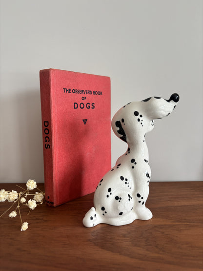 A white ceramic figurine of a Dalmatian dog with black spots sits on a wooden table next to a red hardcover book titled 'The Observer's Book of Dogs' against a plain light gray wall.