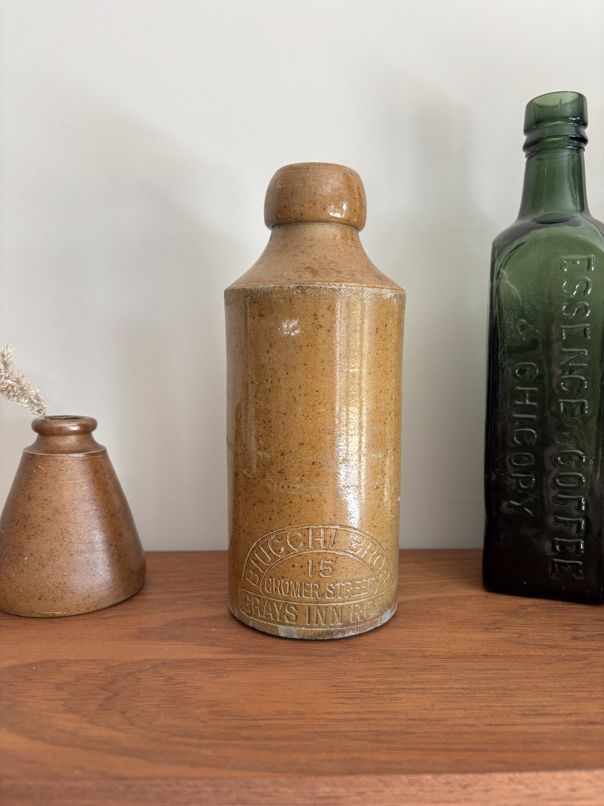 A vintage stoneware bottle with a warm tan-brown speckled glaze and embossed text near the base, displayed on a wooden shelf next to a dark green glass bottle and a small terracotta vase.