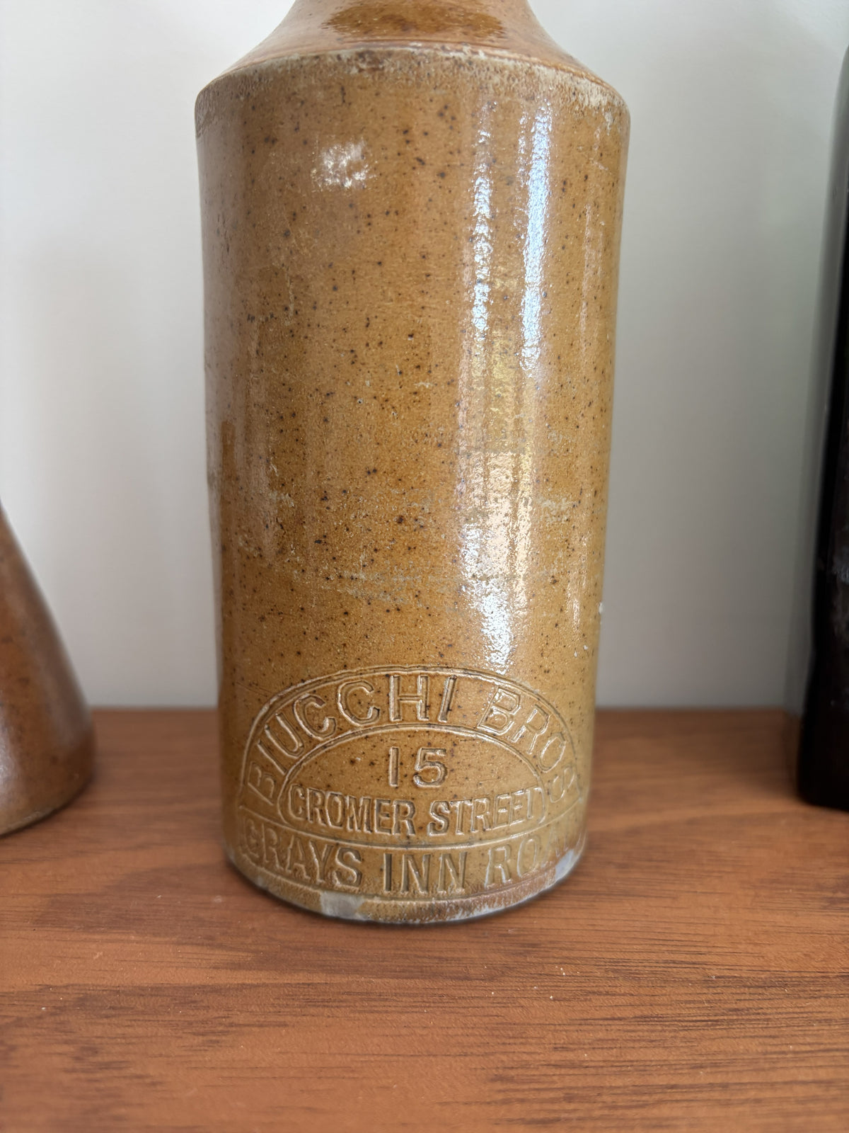 A vintage stoneware bottle with a speckled brown and beige glaze, featuring embossed text that reads 'BIUCCHI BROS' and '15 CROMER STREET GRAYS INN ROAD', displayed on a wooden surface.
