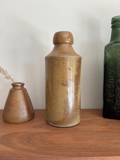 A vintage stoneware bottle with a brown speckled glaze and a matching lid, displayed on a wooden shelf next to a small matching vase and a dark green glass bottle.