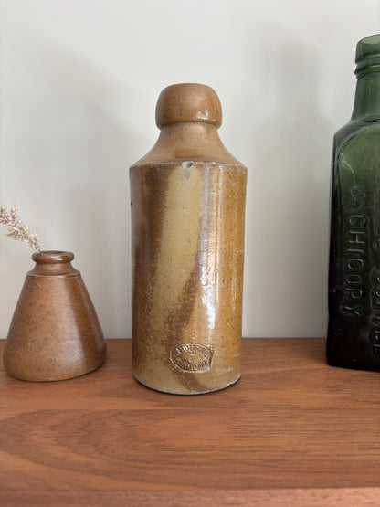 A vintage stoneware bottle with a rounded shoulder and a matching stopper, featuring a brown and beige speckled glaze, displayed on a wooden shelf next to other decorative vessels.
