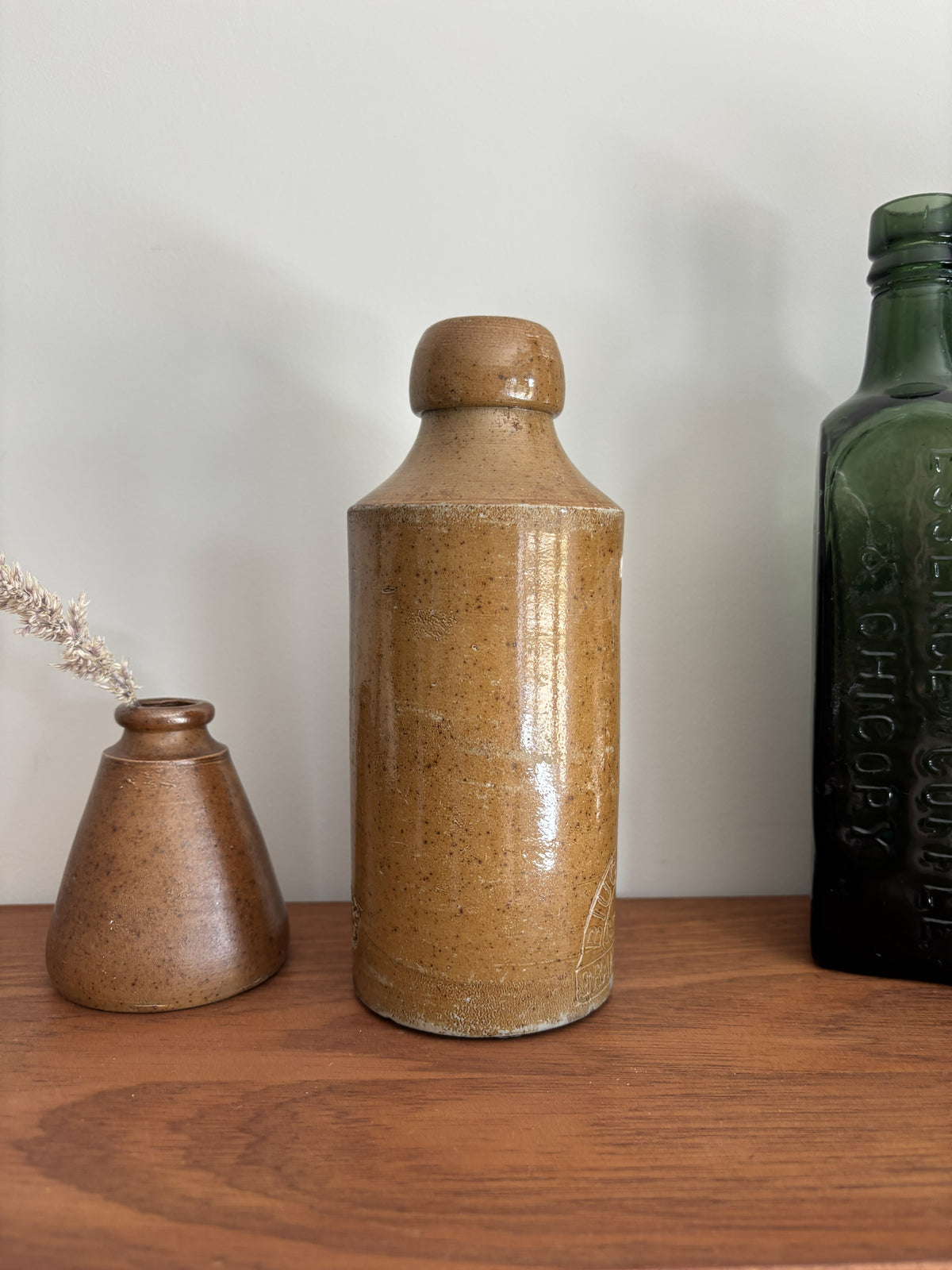 A vintage stone stone bottle with a rounded stopper, displayed on a wooden surface next to a small bronze vase and a dark green glass bottle against a plain light gray wall.