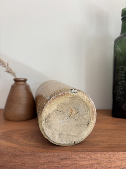 The bottom of a vintage stone bottle with a distressed, chipped glaze lying on its side on a wooden shelf, flanked by a small brown vase and a dark green glass bottle against a plain off-white wall.