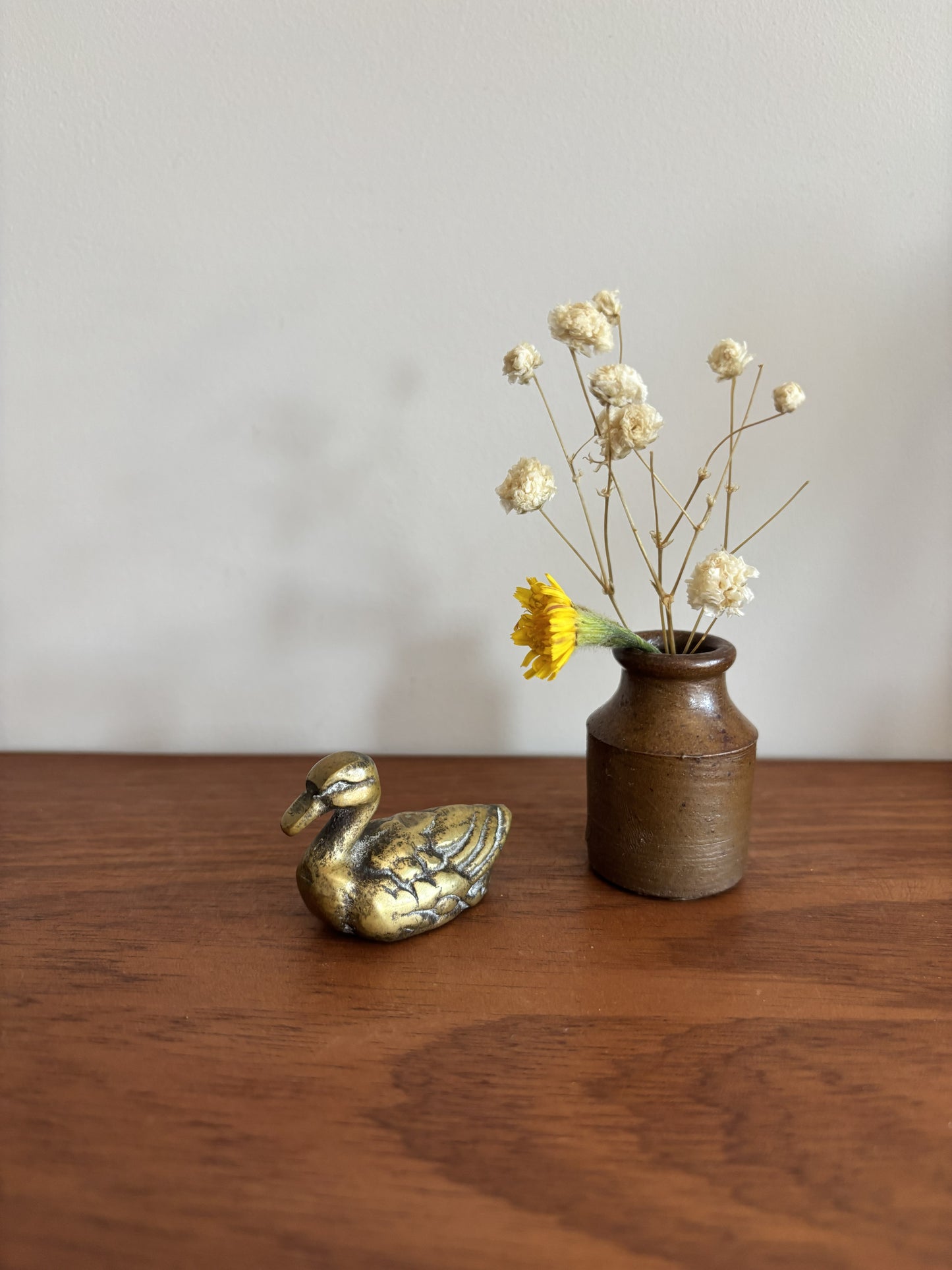 A small brass duck figurine next to a rustic brown ceramic vase with dried white flowers and a yellow accent flower, placed on a wooden table against a plain light gray wall.