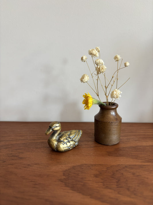 A small brass duck figurine next to a rustic brown ceramic vase with dried white flowers and a yellow accent flower, placed on a wooden table against a plain light gray wall.