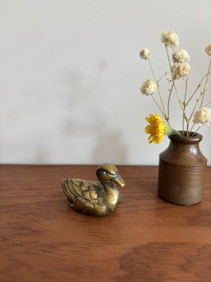 A small brass duck figurine next to a rustic brown ceramic vase with dried white flowers and a yellow accent flower, placed on a wooden table against a plain light gray wall.