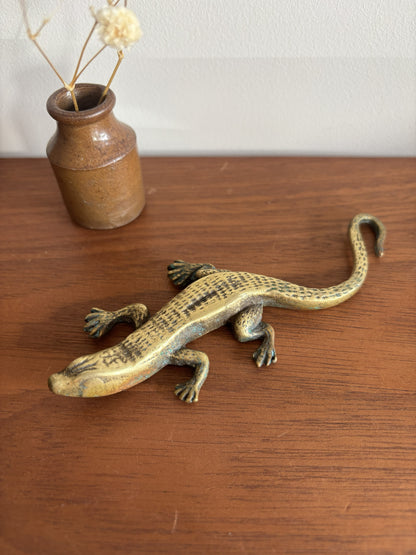 A small brass lizard figurine resting on a wooden tabletop, with a small ceramic vase containing dried stems in the background.