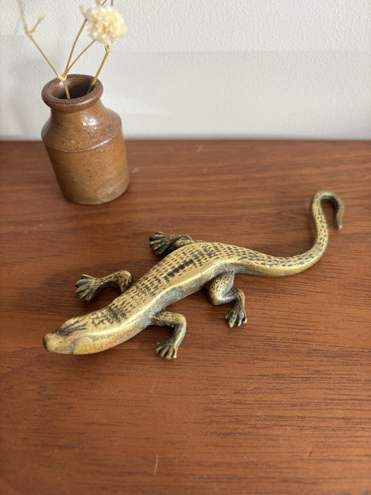 A small brass lizard figurine resting on a wooden tabletop, with a small ceramic vase containing dried stems in the background.