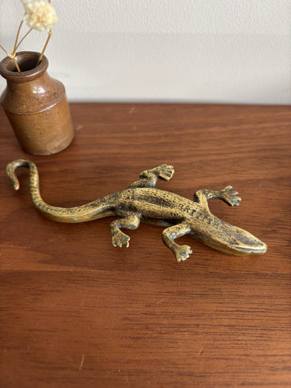 A small brass lizard figurine resting on a wooden tabletop, with a small ceramic vase containing dried stems in the background.