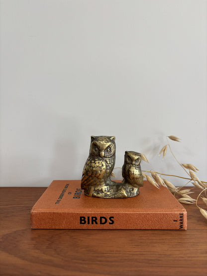 An owl and owlet figurine in a distressed metallic gold and bronze finish, displayed on top of a vintage orange book on a wooden table.