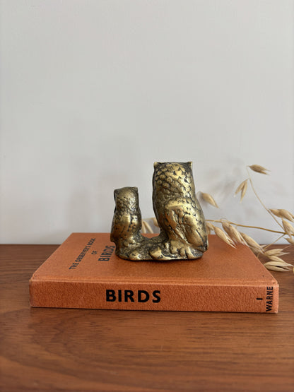 The back of an owl and owlet figurine in a distressed metallic gold and bronze finish, displayed on top of a vintage orange book on a wooden table.