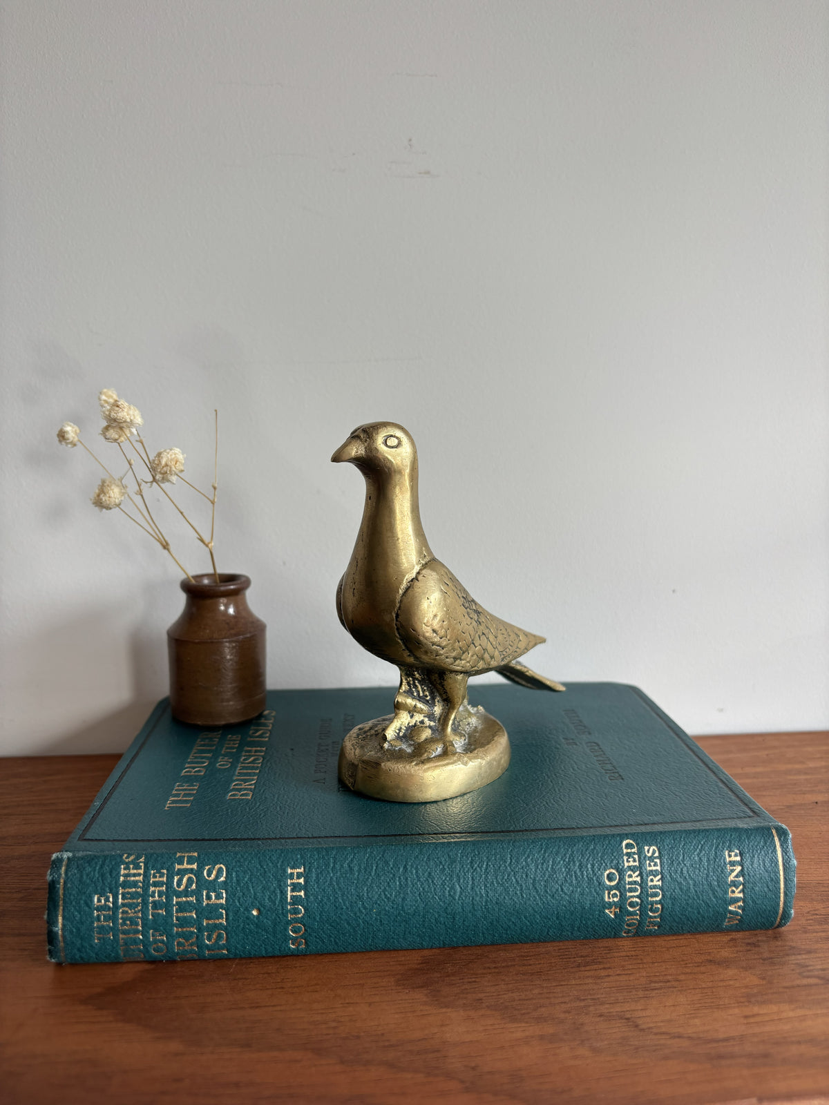 A brass dove or pigeon figurine standing on a round base, placed on top of a teal book. A small brown vase with dried stems sits beside it on a wooden surface against a plain light wall.