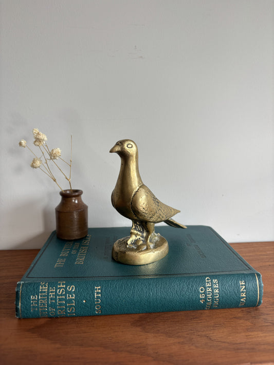 A brass dove or pigeon figurine standing on a round base, placed on top of a teal book. A small brown vase with dried stems sits beside it on a wooden surface against a plain light wall.