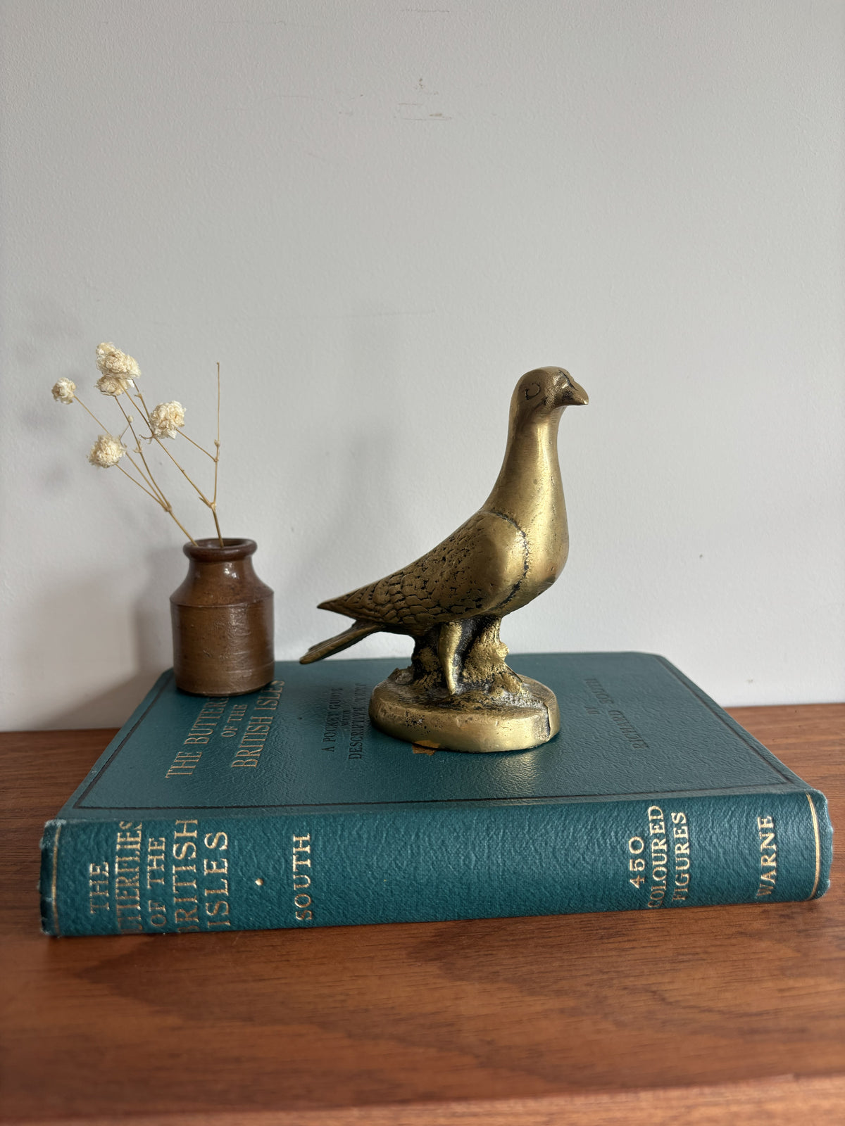 A brass dove or pigeon figurine standing on a round base, placed on top of a teal book. A small brown vase with dried stems sits beside it on a wooden surface against a plain light wall.