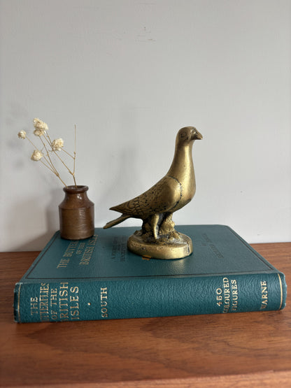 A brass dove or pigeon figurine standing on a round base, placed on top of a teal book. A small brown vase with dried stems sits beside it on a wooden surface against a plain light wall.