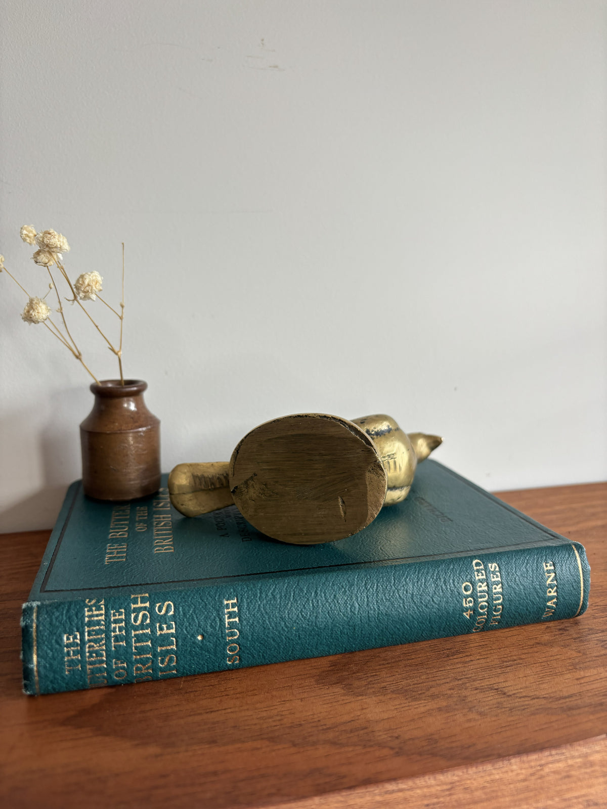 A brass bird figurine lies on its side on top of a vintage teal-blue book, with a small brown vase of dried flowers beside it on a wooden surface against a plain light wall.