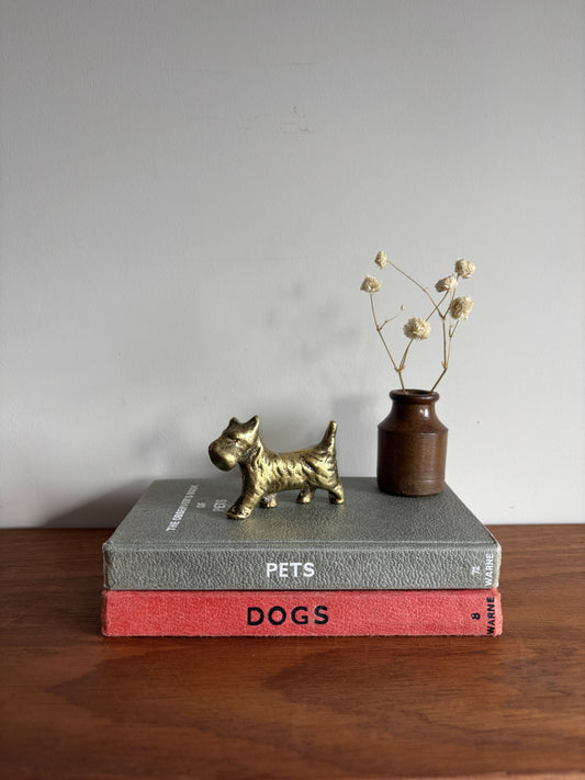 A small brass Scottish Terrier figurine stands on top of two stacked books, one gray and one red, with a small brown vase of dried stems beside it on a wooden surface against a plain light gray wall.